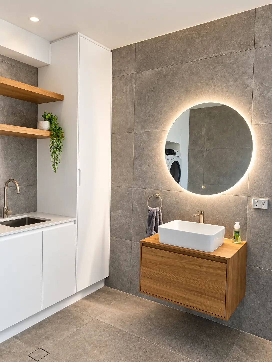 Grey tiled bathroom with illuminated round mirror, timber vanity and integrated laundry joinery